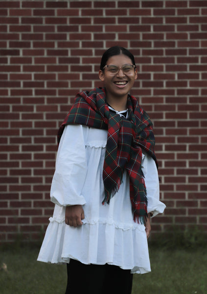 Person wearing a white dress and Quebec tartan red, green and white plaid scarf in front of a brick wall