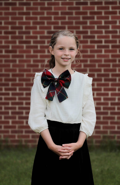 Young girl in a white blouse with a Quebec tartan bow tie and black skirt standing in front of a brick wall.