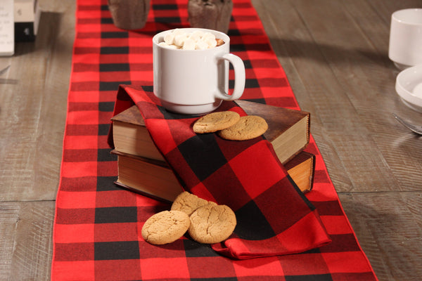 Close-up of a Queen Charlotte's Check plaid napkin on a stack of books, topped with a cup of hot chocolate and cookies, with a matching table runner underneath placed on a wooden table.