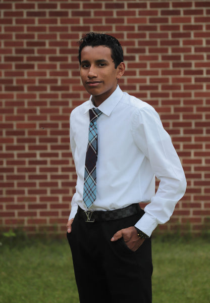 Young man wearing a blue and maroon plaid necktie with a white shirt and black dress pants standing in front of a brick wall.
