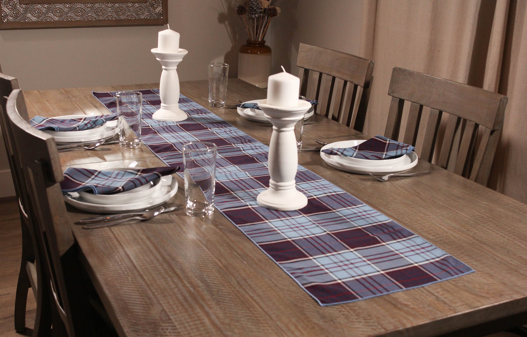 Blue and mauve tartan table runner with matching napkins displayed on a wooden table, set with white plates and white candles.