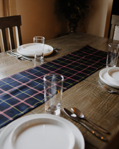 A RCN tartan navy blue table runner with a red and yellow plaid pattern laid out on a rustic wooden dining table, surrounded by white plates, clear drinking glasses, and silver cutlery.