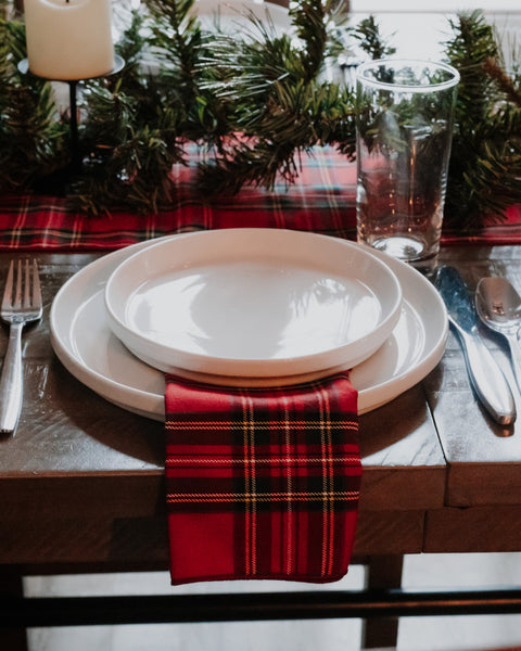 A festive holiday table setting featuring a red and black plaid napkin under stacked white ceramic plates, with a matching plaid table runner, pine garland, glassware, and silver cutlery on a dark wooden table.