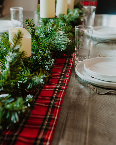 A festive holiday table runner made of Royal Stewart tartan red plaid fabric and green pine garland, styled on a wooden dining table with white plates, clear drinking glasses, and white pillar candles.