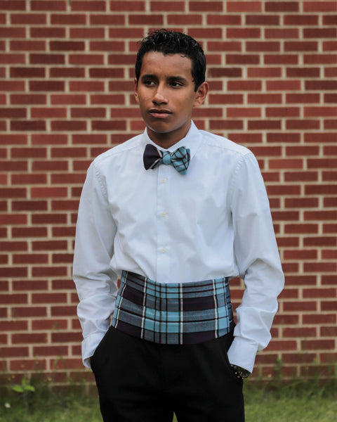 Person wearing a white shirt, plaid cummerbund and  RCAF plaidbow tie in front of a brick wall.