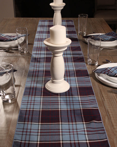 Dining table set with a blue and plum plaid runner, white candlesticks, and glasses.