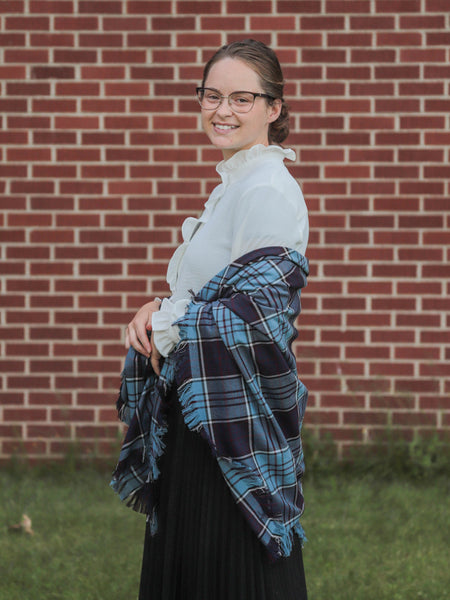 Woman wearing a white blouse and Air Force plaid shawl against a brick wall.