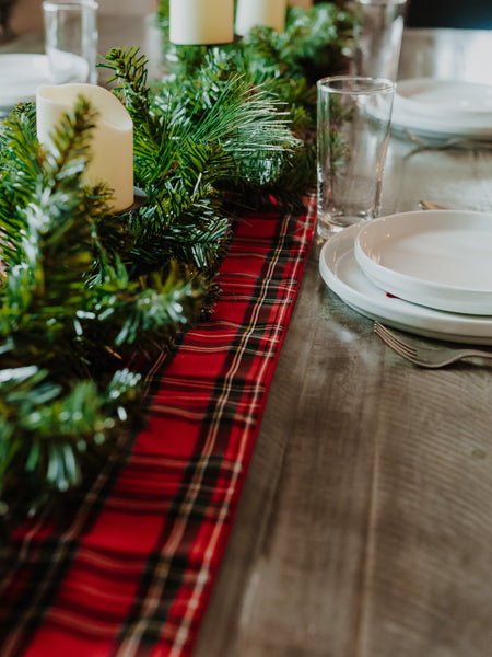 A festive holiday table runner made of red, black, and green plaid fabric, styled with pine greenery, white pillar candles, and white plates on a rustic wooden table.
