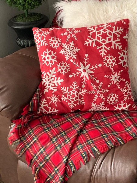 Red pillow with white snowflake pattern on  red plaid blanket a brown couch