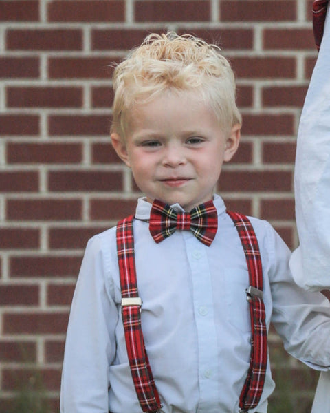 Young boy wearing a red plaid bow tie and suspenders against a brick wall.