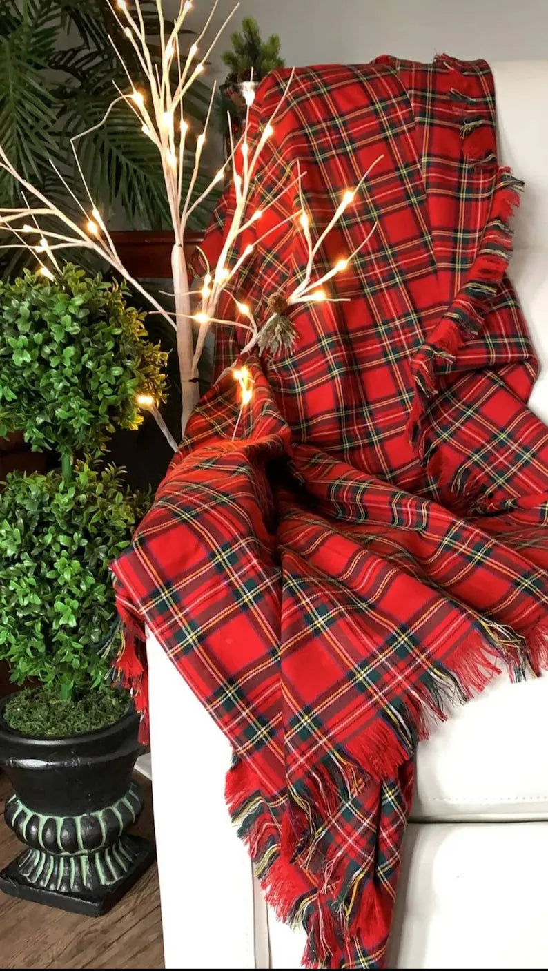 A red and green plaid Royal Stewart tartan throw draped over a white chair with Christmas decorations in the background, including a tree and fairy lights.
