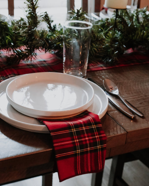 A festive holiday table setting featuring a red and black plaid Royal Stewart tartan  napkin, white ceramic plates, silver cutlery, and a clear drinking glass, all arranged on a rustic wooden table with a pine garland runner in the background.