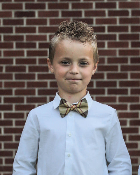 Young boy wearing a white shirt and a tan gold green and red patterned bow tie against a brick wall.