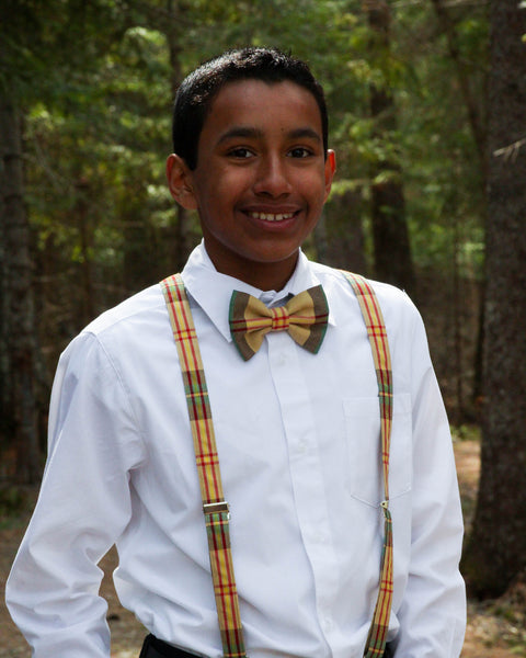 Young boy wearing a white shirt, yellow plaid bow tie, and suspenders standing in a forest.