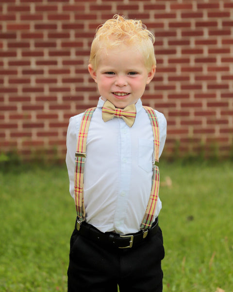 Smiling blonnd boy wearing a yellow plaid bow tie and suspenders with his hands behind his back. 
