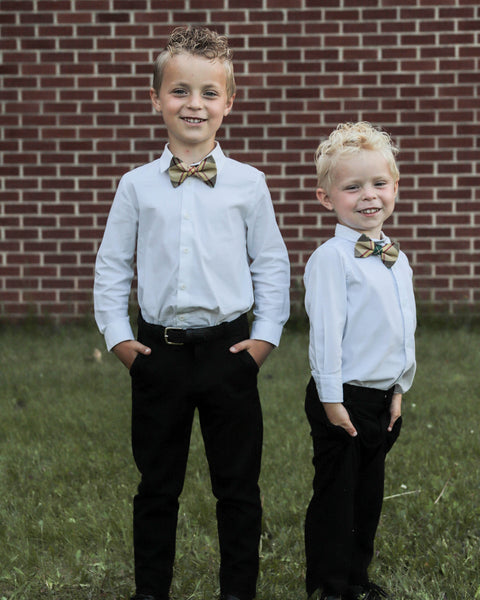 Two young boys in white shirts and black pants with bow ties standing in front of a brick wall.