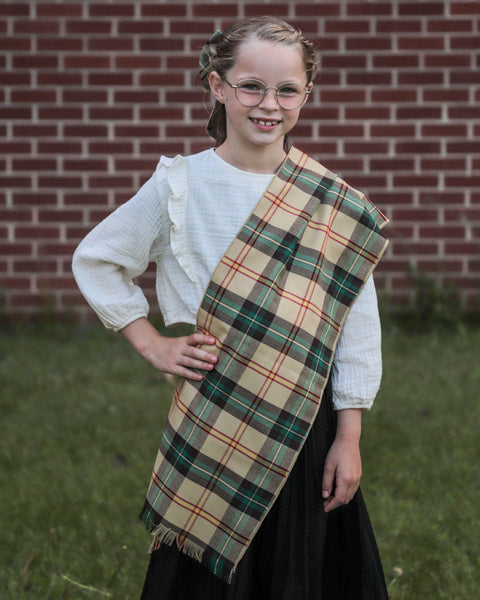 Young girl wearing a yellos plaid sash in front of a brick wall.