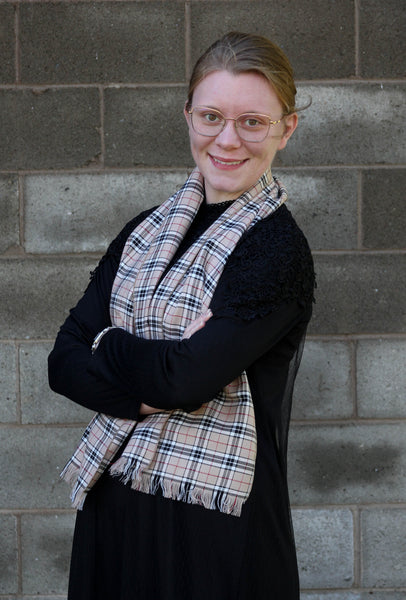 Woman wearing a black top and Thompson tartan scarf, standing with arms crossed in front of a brick wall.