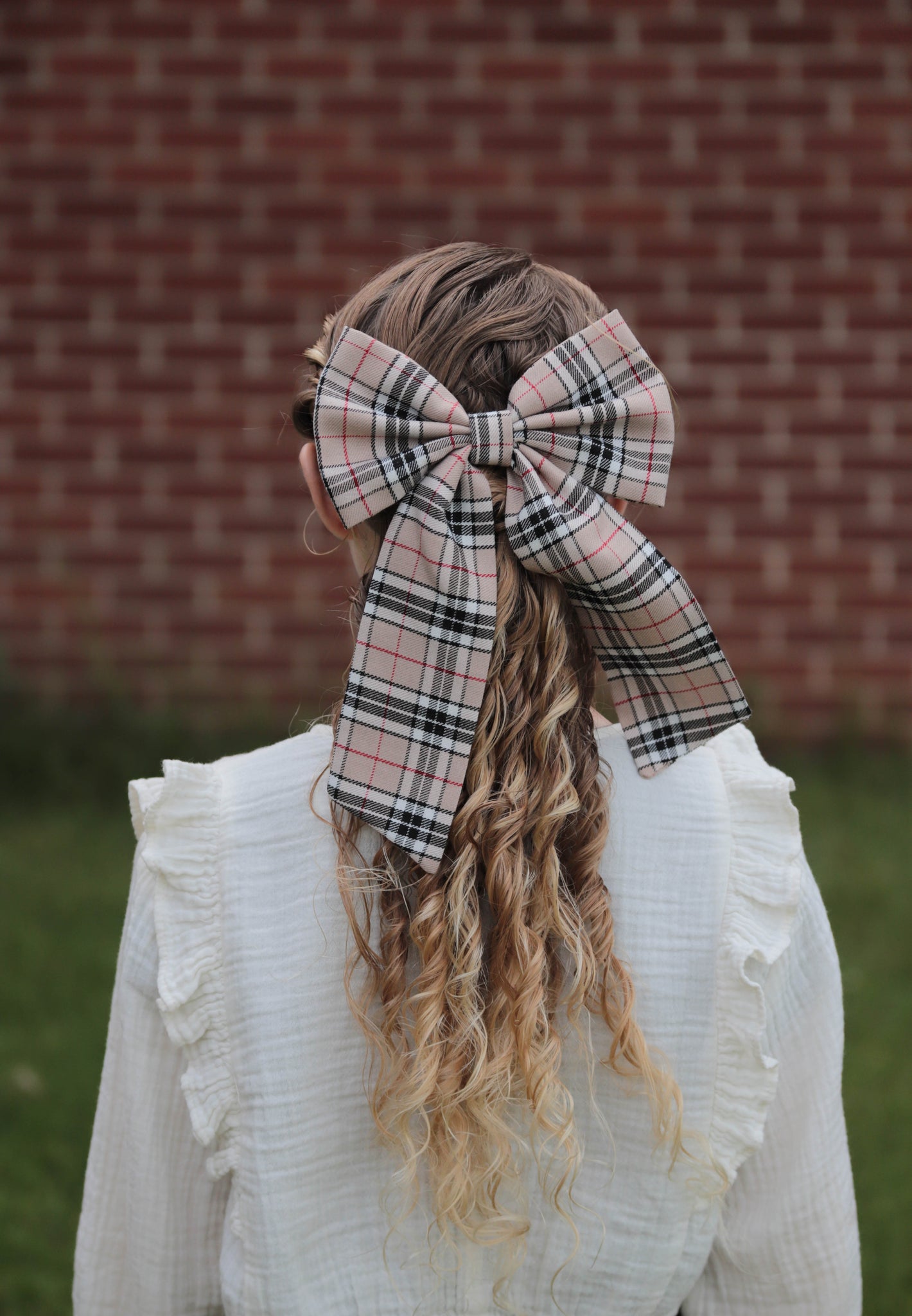 Girl with blonde curly hair wearing a white blouse and a tan, black, and white Thompson tartan hair bow.