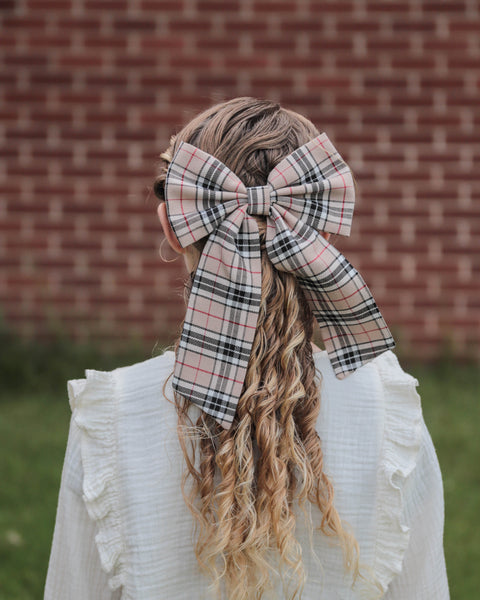 Girl with blond curly hari wearing tan, black, white and red plaid oversize bow tie with white ruffle blouse looking at brick wall