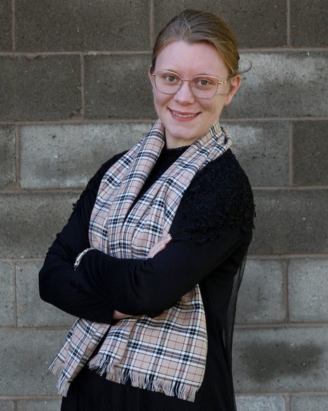 Woman wearing a Thompson plaid scarf over black dress and glasses against a brick wall