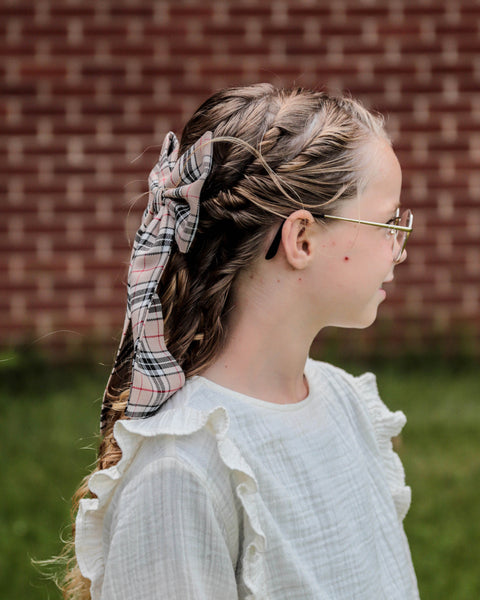 Girl looking away wearing tan, white and blackThompson tartan hair bow in her curly hair, outdoors, brick wall and green grass behind.