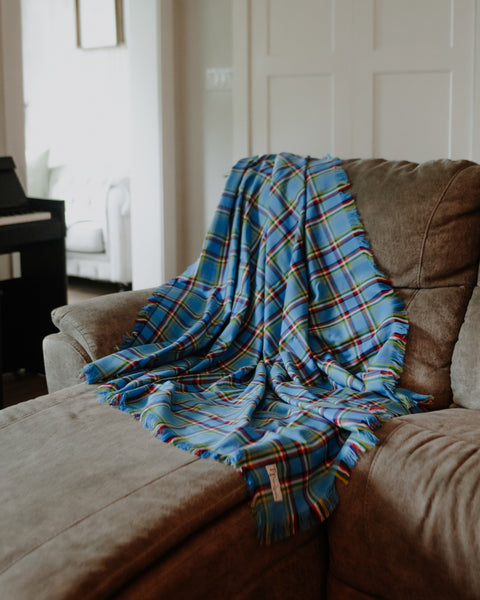 A Yukon tartan blue plaid throw blanket with multicolor stripes and fringe is draped over a brown fabric sofa in a living room setting.