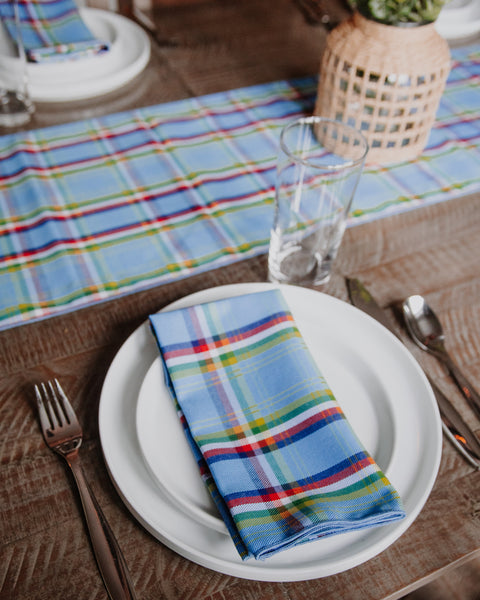 A folded blue plaid Yukon tartan napkin with red, green, and yellow stripes is displayed on a white plate on a rustic wooden table, alongside a glass, fork, and spoon.