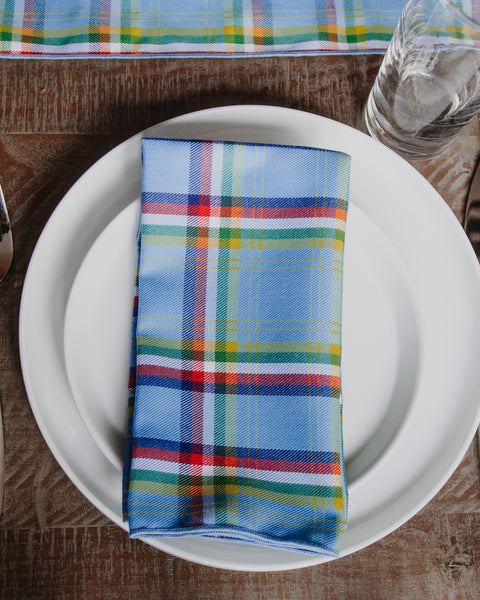A folded plaid napkin in light blue with red, green, yellow, and white stripes, placed on a white plate on a rustic wooden table.