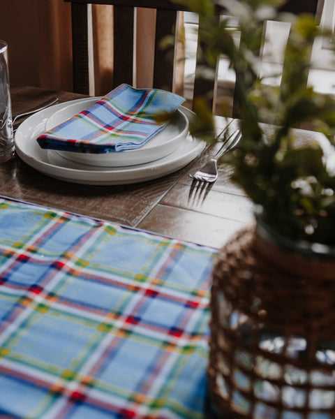A blue plaid Yukon tartan tablecloth and matching napkin set on a wooden dining table, styled with white plates, a fork, and a potted plant in the foreground.