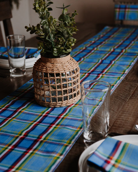A Yukon tartan blue plaid table runner with red, green, and yellow stripes laid across a wooden dining table, styled with a woven basket vase holding green foliage, clear drinking glasses, and white plates.