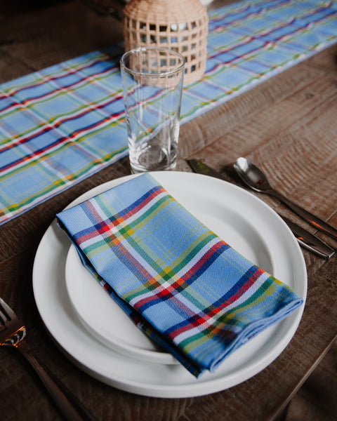 A folded Yukon tartan blue plaid napkin with red, yellow, and green stripes is displayed on a white plate on a rustic wooden table, alongside a clear drinking glass, silver cutlery, and a matching Yukon plaid table runner.