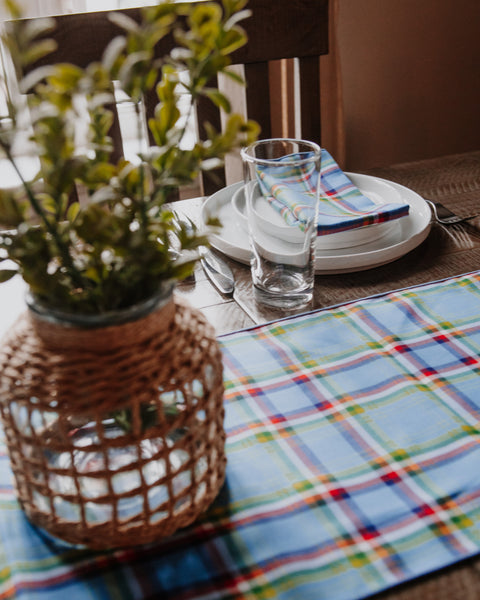 A blue plaid table runner with red, yellow, and green stripes laid on a wooden dining table, styled with white plates, a clear drinking glass, a folded striped napkin, and a woven basket planter in the foreground.