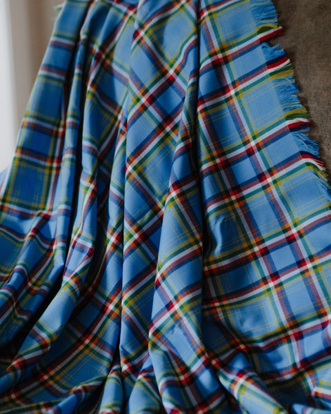 A close-up of a draped blue plaid fabric with fringed edges, showing a tartan pattern in blue, red, yellow, and white.