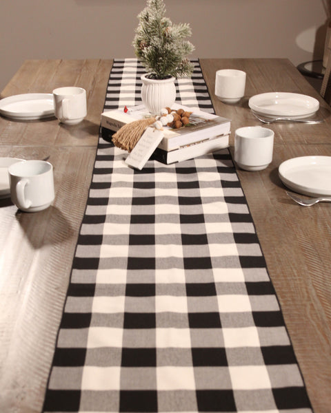 Dining table set with a black and white checkered table runner, white plates, cups, and a small plant.