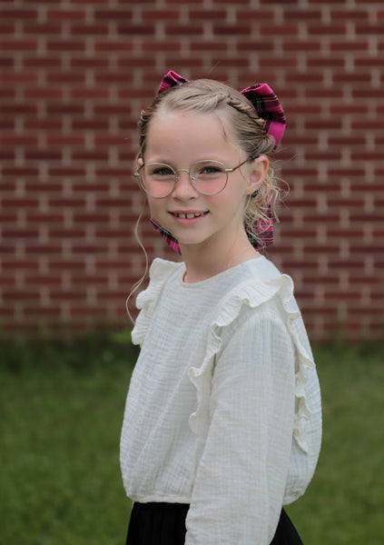 Young girl with glasses in a white ruffled sweater and pink bow, standing in front of a brick wall.