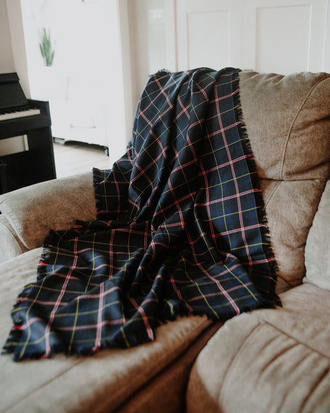 A Royal Canadian Navy tartan dark navy blue plaid throw blanket with pink, yellow, and white stripes and fringed edges, draped over a beige fabric armchair in a living room setting.