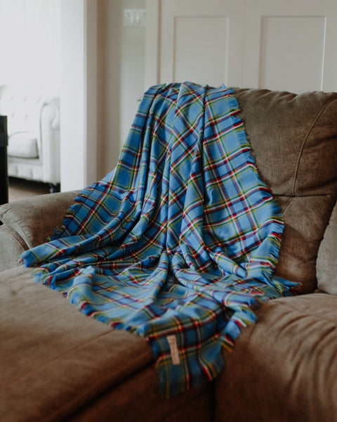 A Yukon tartan blue plaid throw blanket with red, yellow, and green stripes draped over a brown fabric couch in a living room setting.