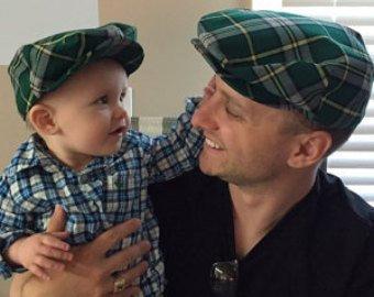 Adult male holding a smiling toddler. They’re looking at each other, both wearing green, yellow, and gray Cape Breton tartan flat caps.